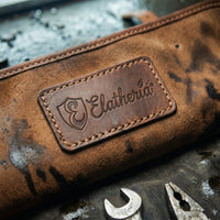 Macro close-up of the embossed leather Elatheria logo patch and heavy-duty contrast stitching on a distressed brown leather tool roll flap.