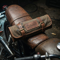 Fully rolled up and buckled Heritage Brown leather tool roll by Elatheria resting on the distressed leather seat of a vintage cafe racer motorcycle.