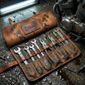Open Elatheria Heritage Brown leather and waxed canvas tool roll lying on a gritty garage floor next to a vintage motorcycle engine, securely holding silver wrenches and pliers.
